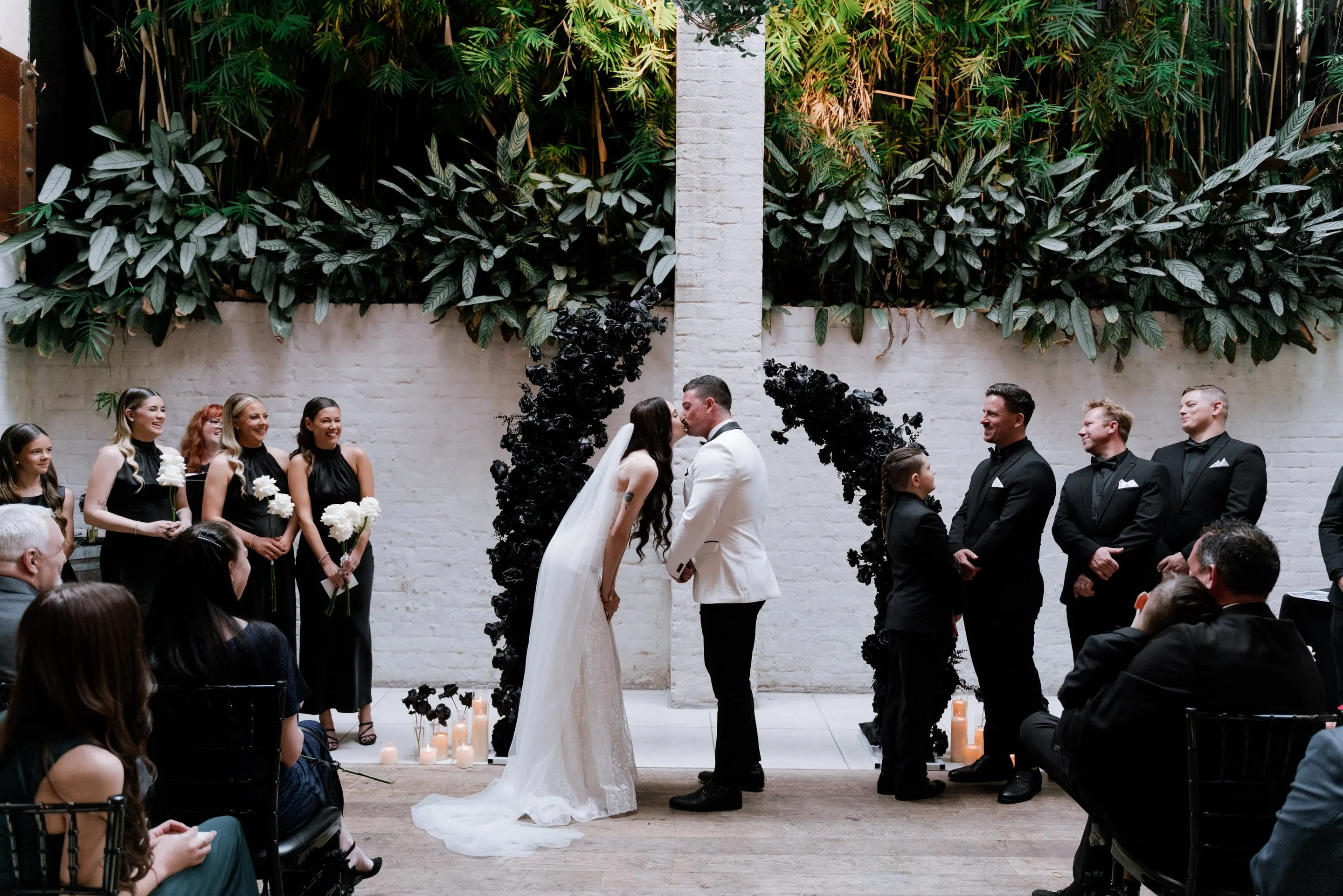 Couple kissing between black floral arches in a courtyard, with their bridal party and guests either side