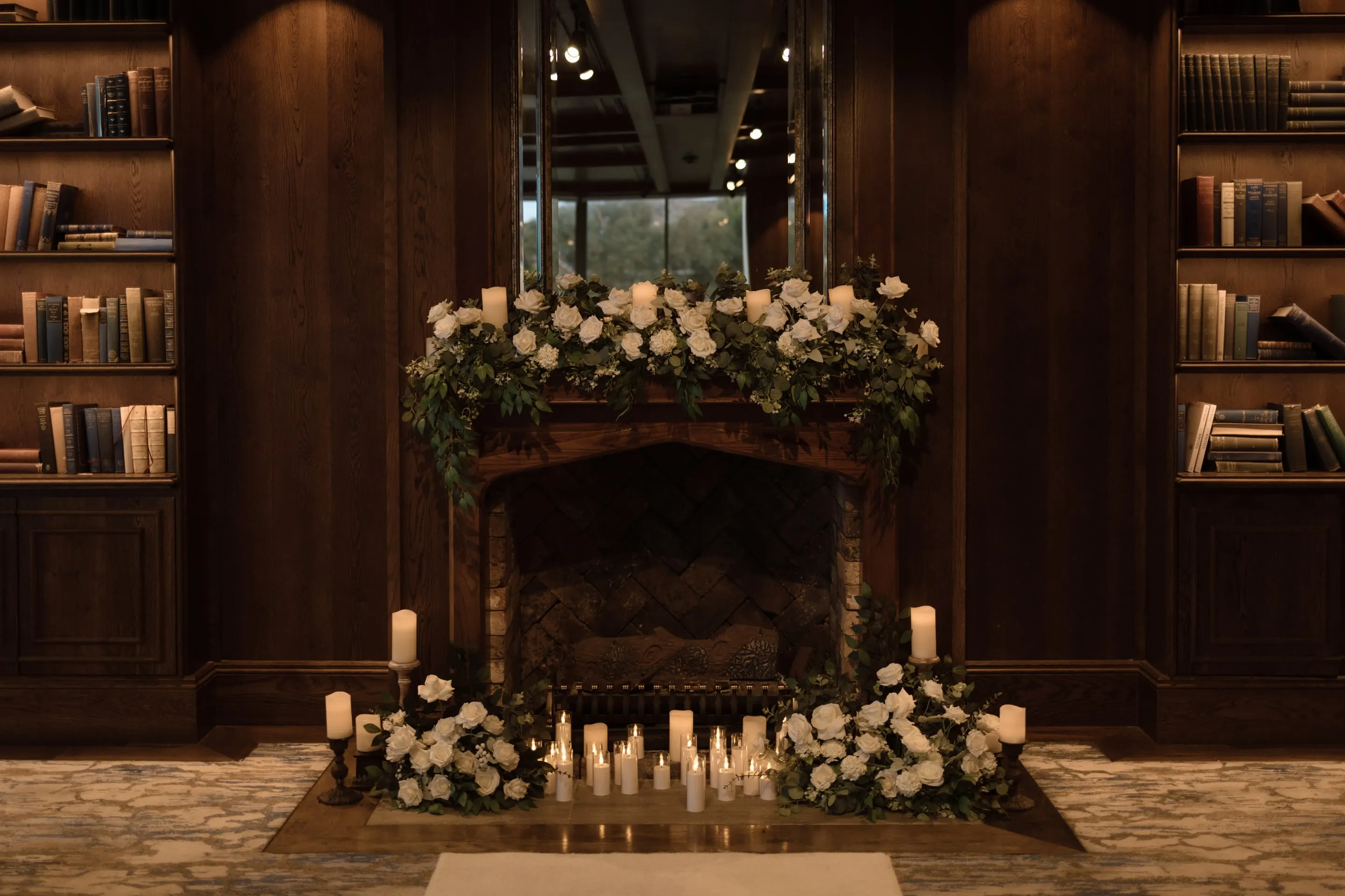 Timber library fireplace dressed with white roses, greenery and pillar candles for an indoor ceremony