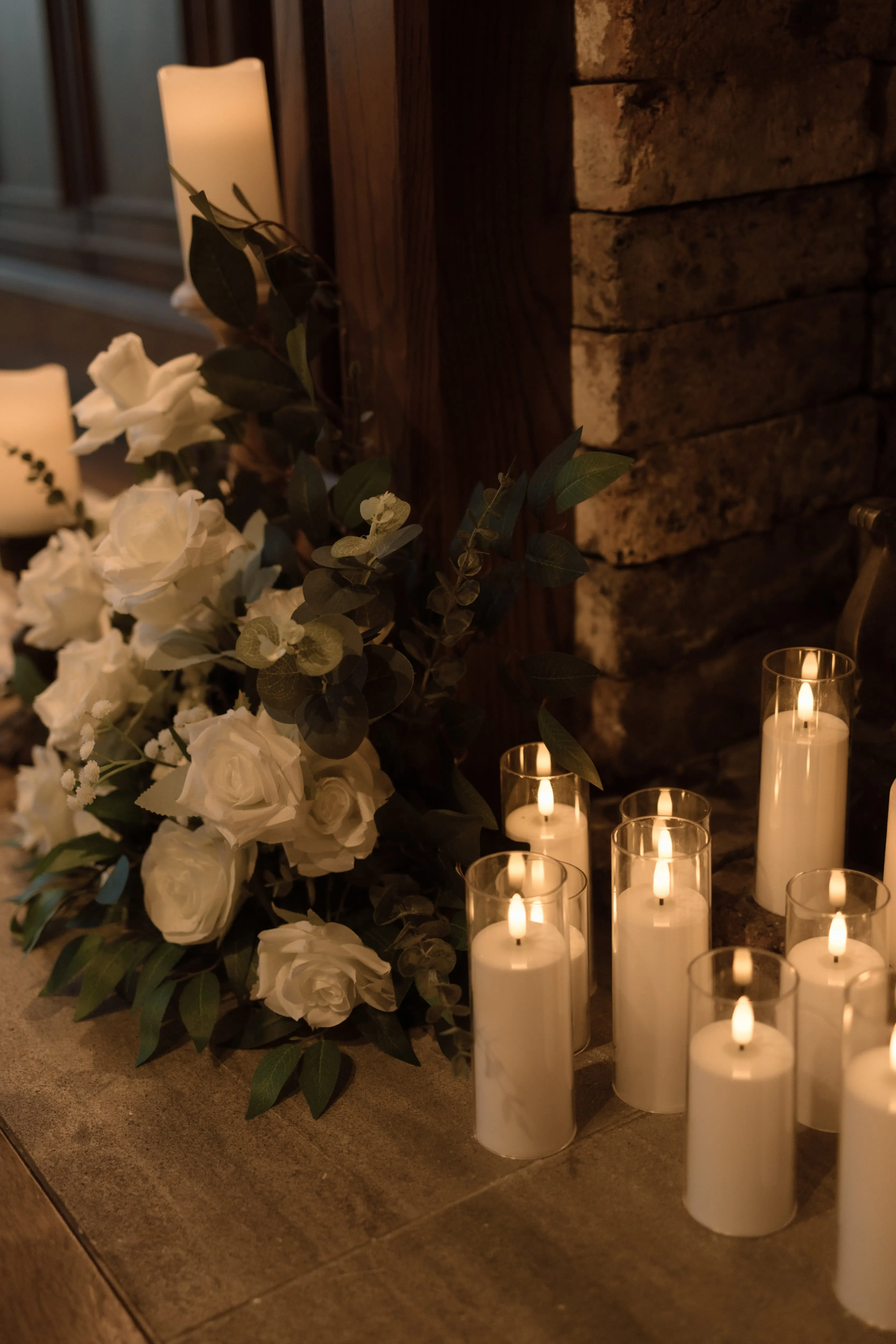Pillar candles glowing in glass holders beside white roses and greenery