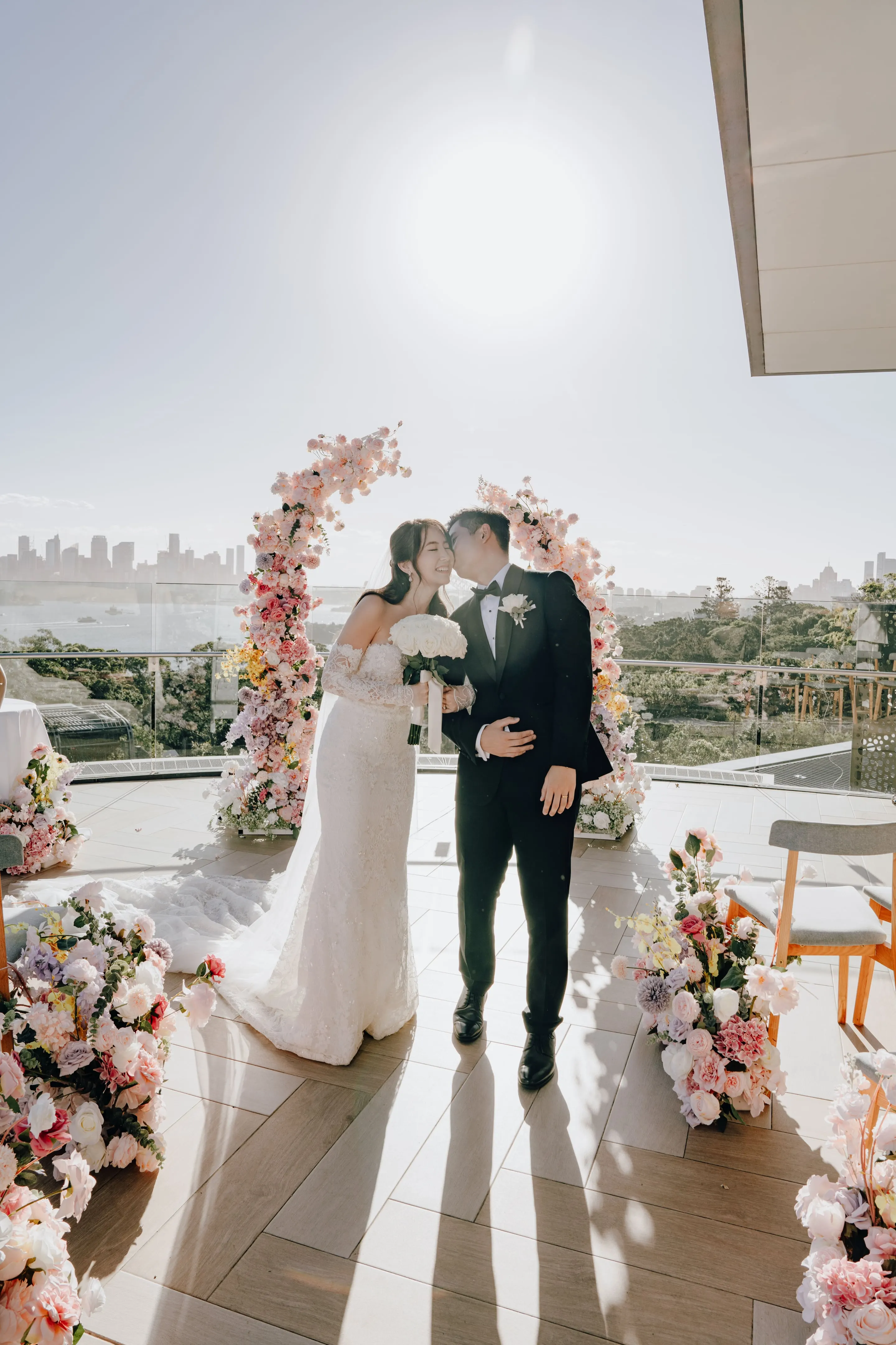 Newlyweds kissing between pastel floral arches on a harbourside terrace, the city skyline behind