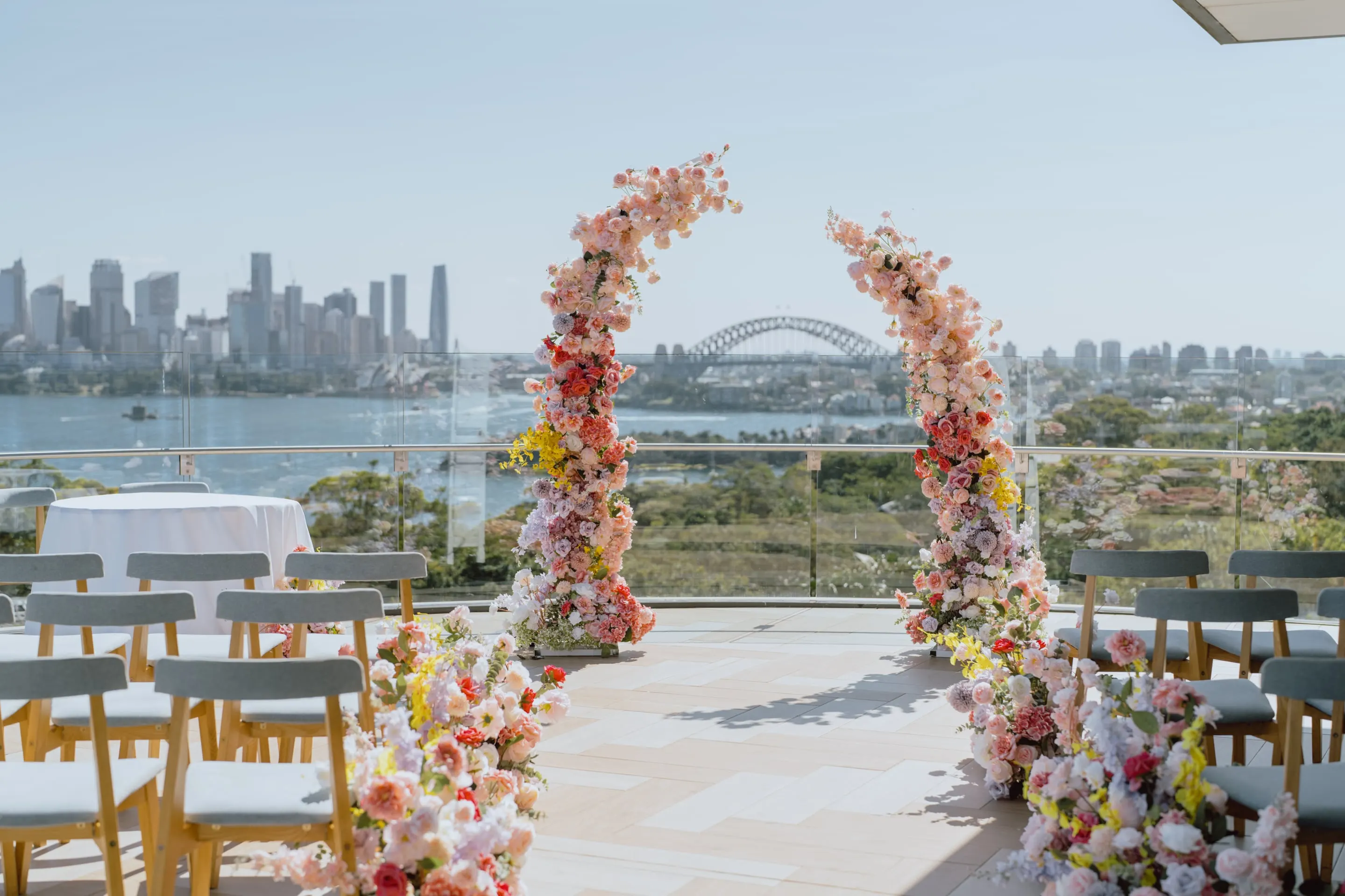 Two floral arches in pink, peach and yellow framing a harbourside ceremony, with the Harbour Bridge and city skyline behind