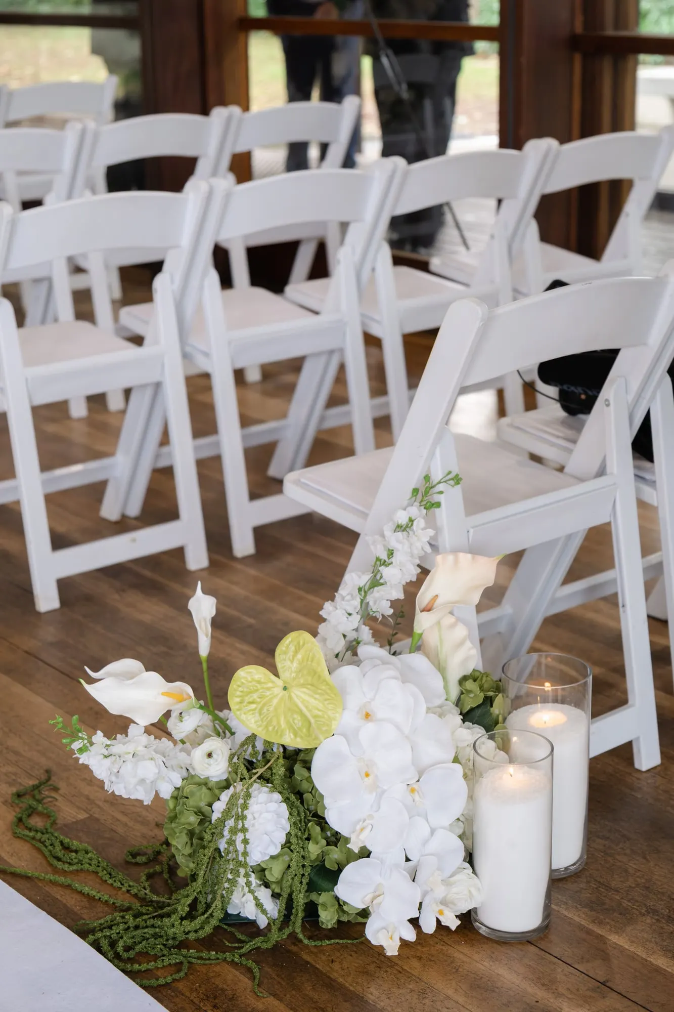 White orchids, calla lilies and pillar candles at the end of a row of white ceremony chairs