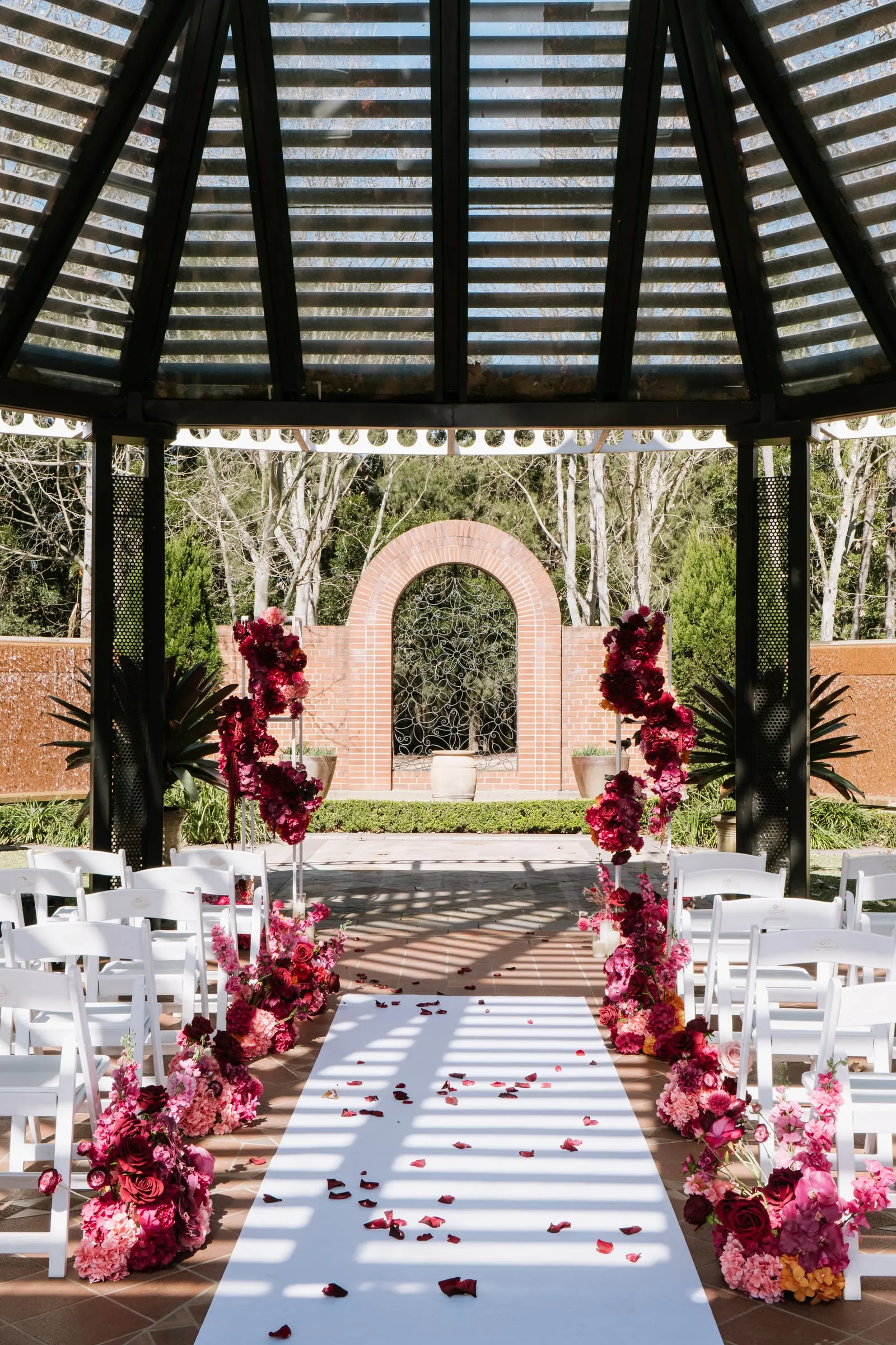 Garden gazebo ceremony with white chairs, a white aisle runner and tall red and pink floral columns