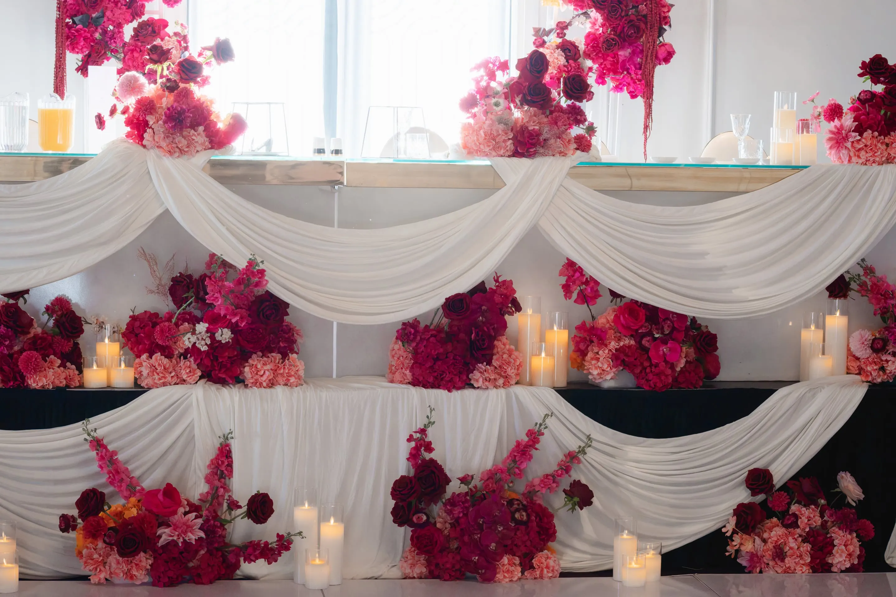 Bridal table dressed in draped white fabric, with red and pink florals and candles on two tiers