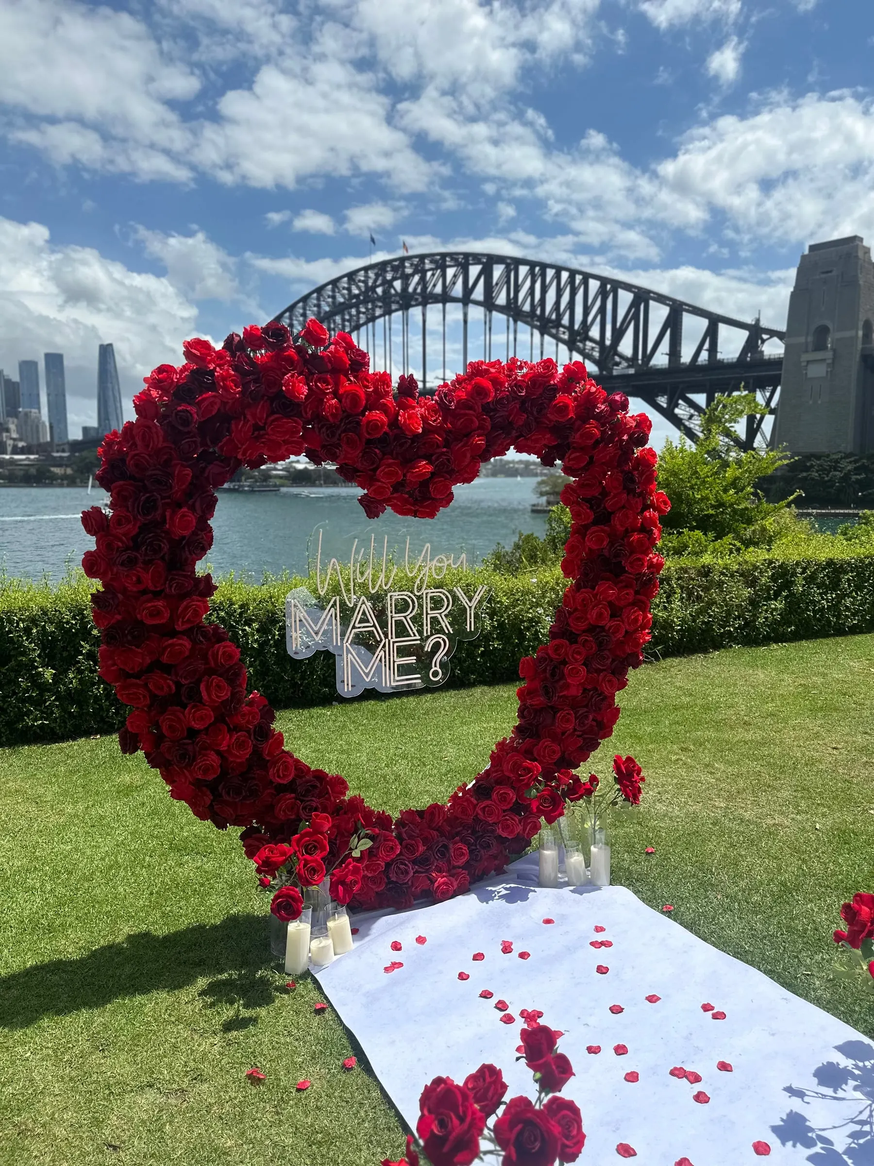 Red rose heart arch with a Marry Me sign and petal-scattered runner, the Harbour Bridge behind
