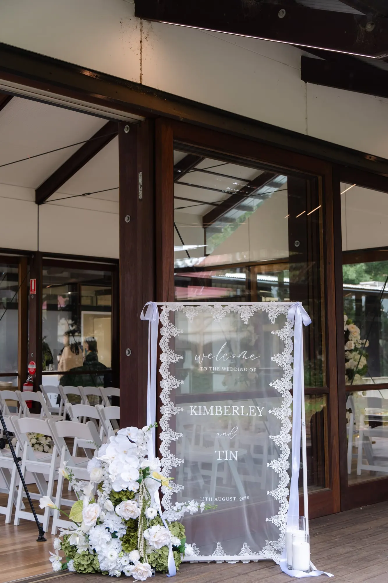 Lace-draped welcome sign with white florals in a timber pavilion