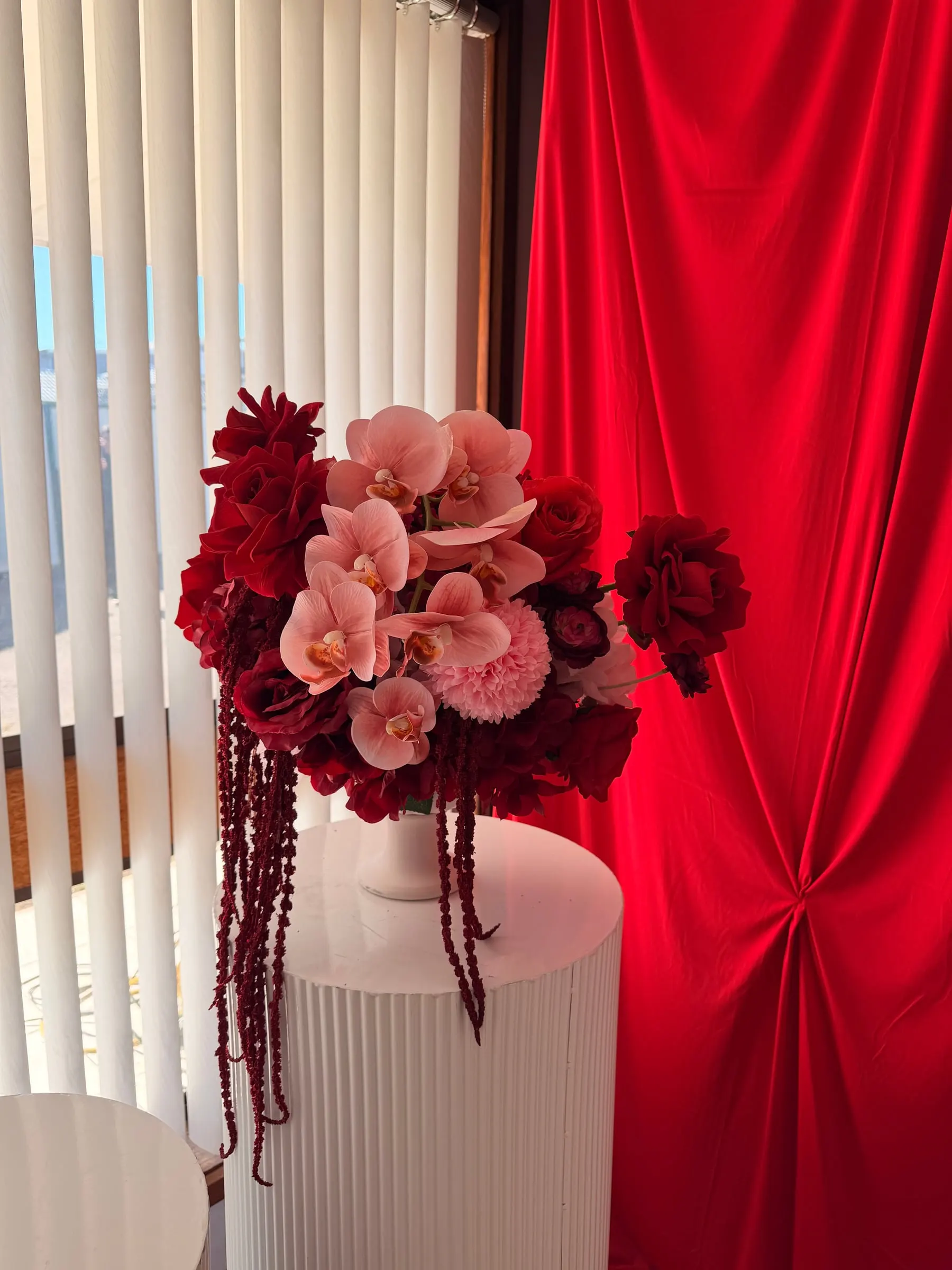 Arrangement of red roses, pink orchids and trailing amaranthus on a white plinth before a red drape