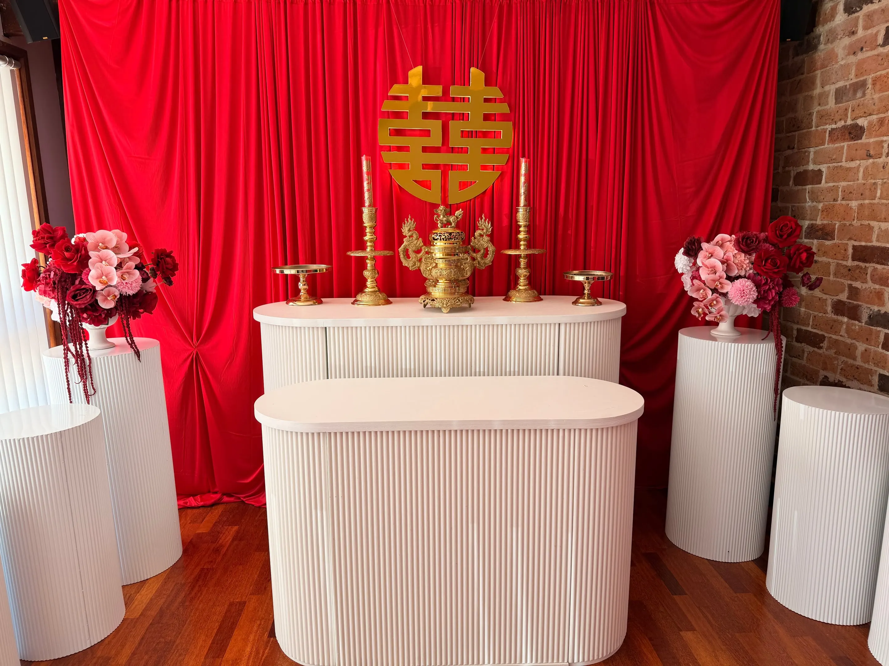 Tea ceremony altar with a gold double happiness sign, gold candlesticks, white fluted tables and pink and red florals before a red drape
