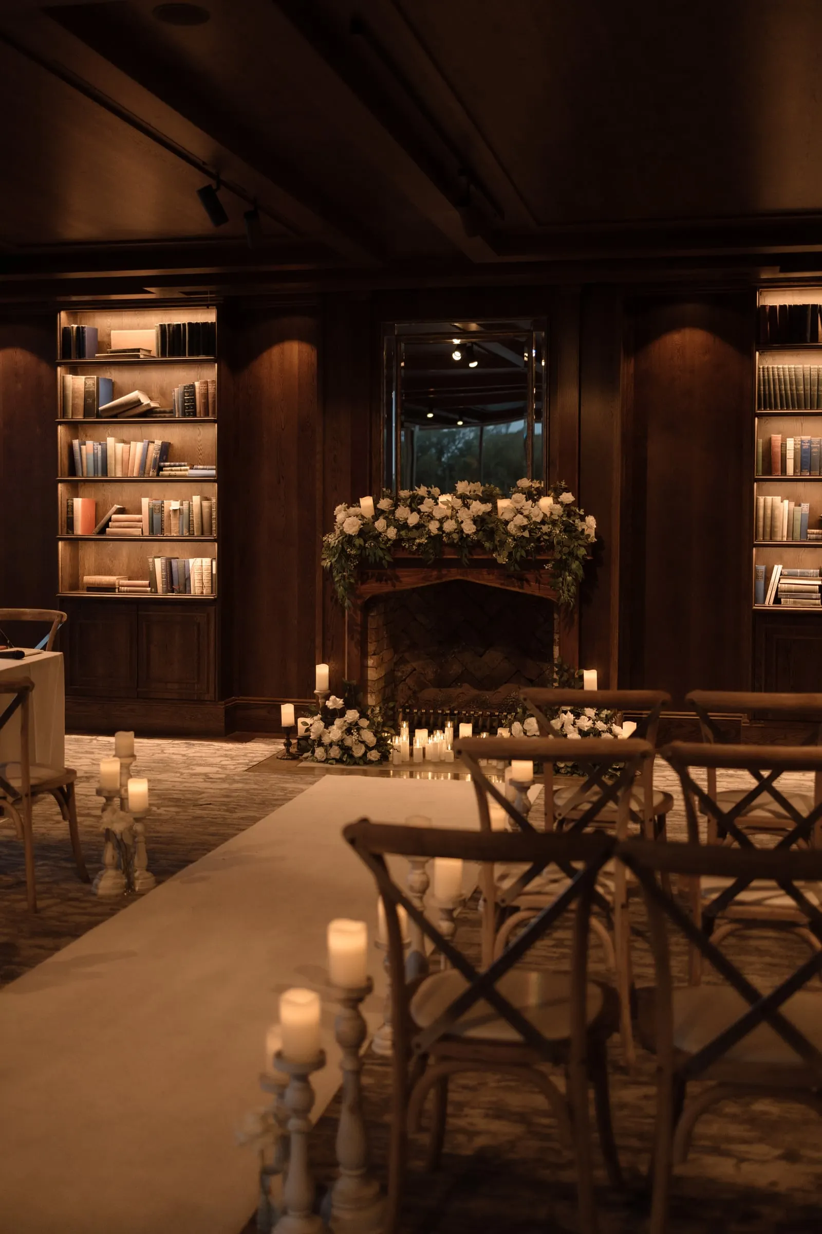 Ceremony aisle lined with pillar candles in glass stands, with florals at the fireplace beyond