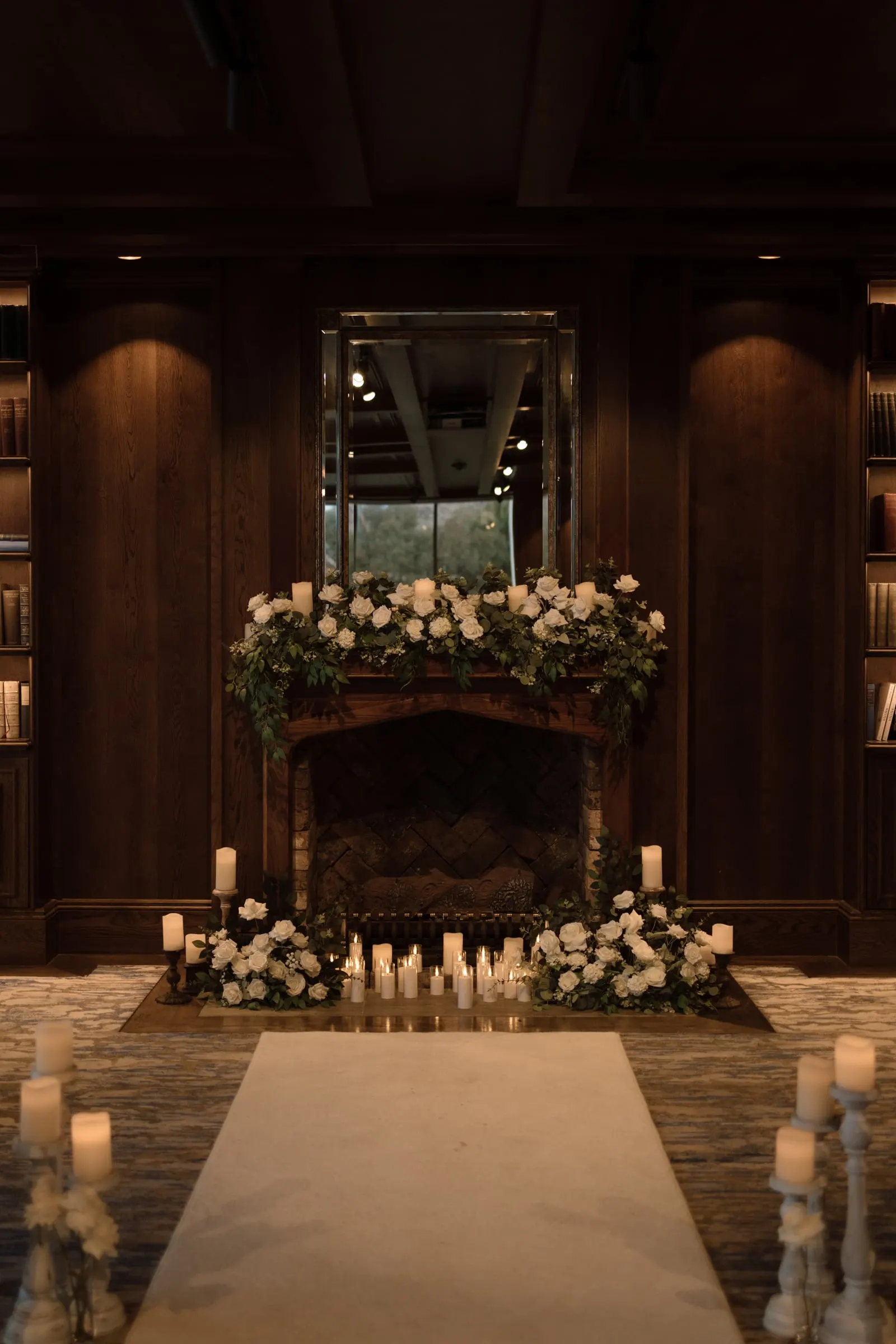 Candlelit aisle leading to a fireplace dressed in white roses in a timber library room
