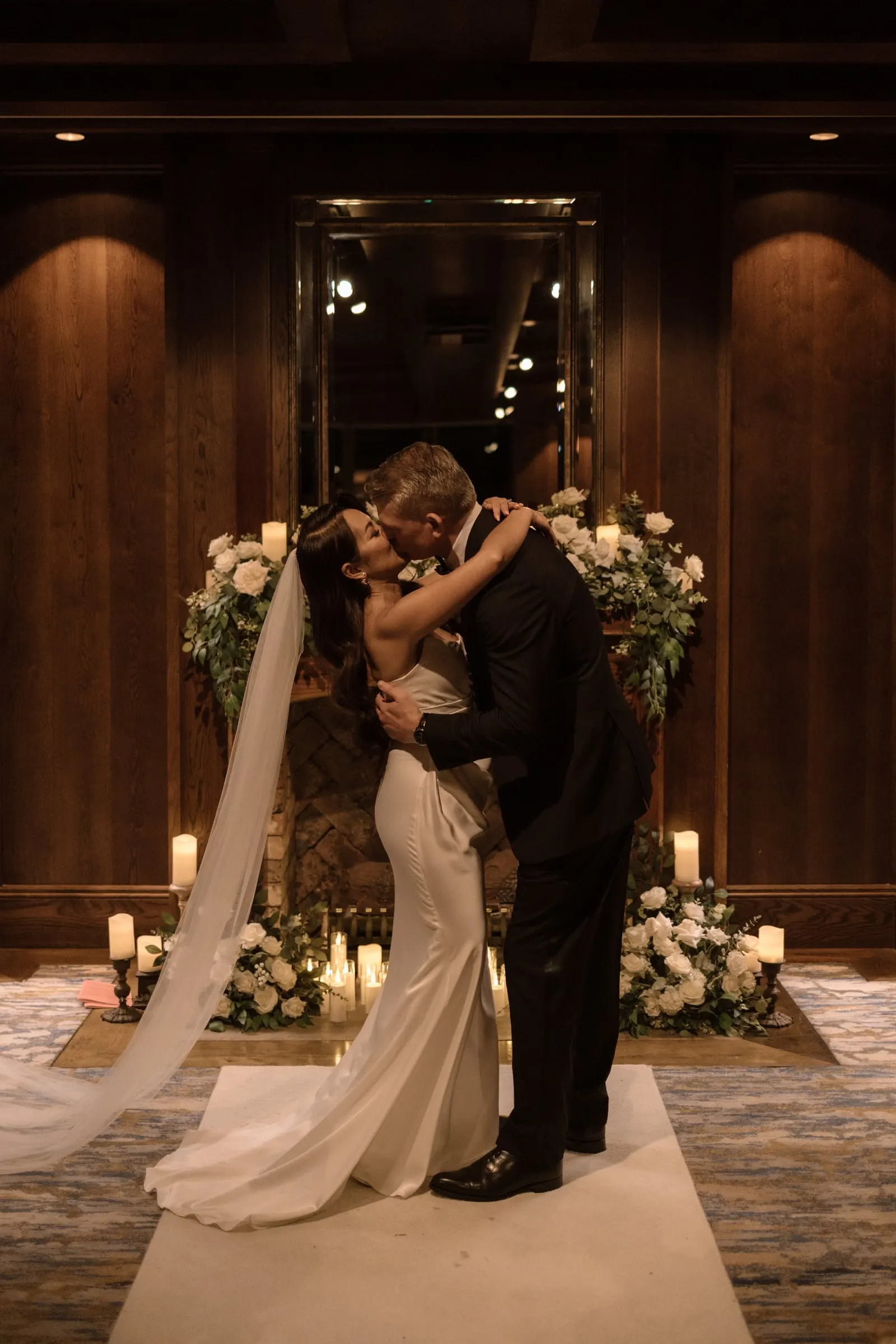 Couple kissing at the end of a candlelit aisle in front of a flower-covered fireplace
